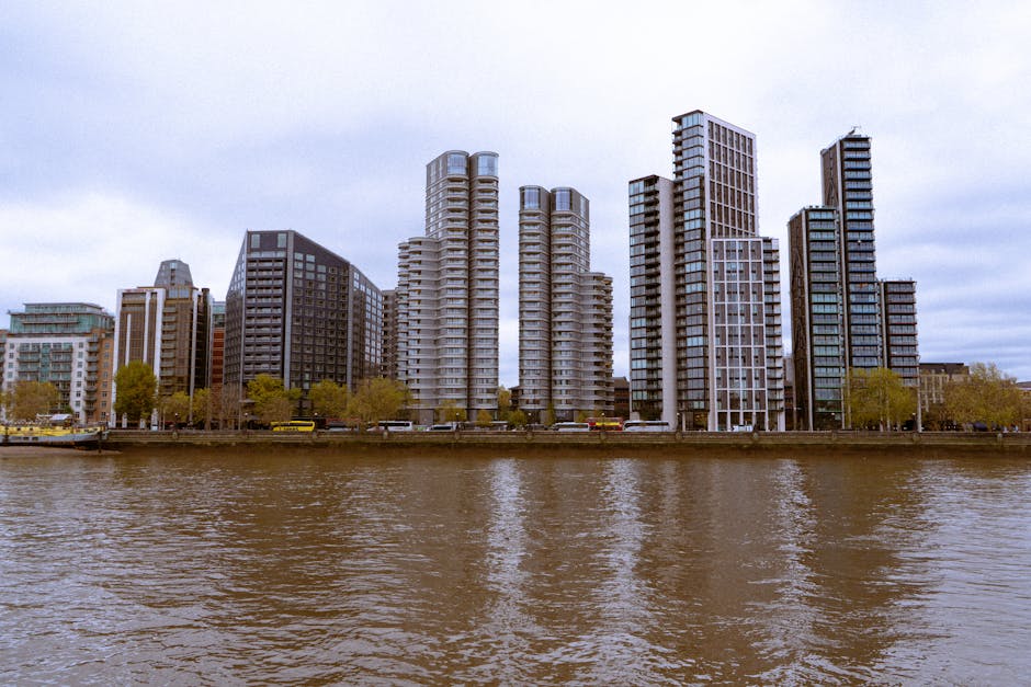 A wide view of a modern city skyline taken from across a river, featuring tall residential and office buildings with glass and concrete facades. The buildings are clustered along the riverbank, with s