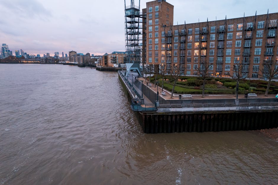 A wide view of a modern city skyline taken from across a river, featuring tall residential and office buildings with glass and concrete facades. The buildings are clustered along the riverbank, with s